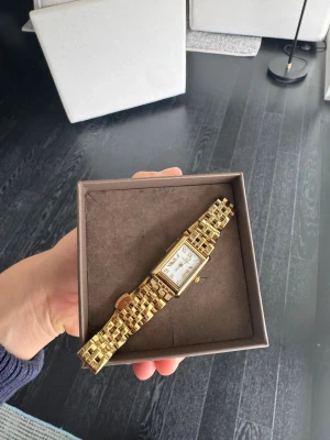 Guldfärgad  klocka - Stilren guldfärgad klocka med rektangulärt ur och länkarmband. Urtavlan är vit med romerska siffror och gulddetaljer. Perfekt accessoar för dig som gillar klassisk och elegant stil.