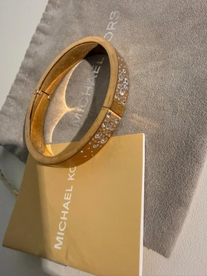 Guldigt armband Michael Kors - Snyggt guldigt armband från Michael Kors med gnistrande stenar på ovansidan. Armbandet har en stilren och elegant design med ett smidigt gångjärn för enkel påtagning. Perfekt accessoar för att lyfta din outfit med lite extra bling.