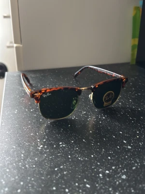 Ray-Ban Clubmaster solglasögon brun - Snygga Ray-Ban Clubmaster solglasögon med klassisk brunsköldpaddsmönstrad båge och gröna glas. Metallbåge i guldton och retroinspirerad form som ger en tidlös look. Perfekta för soliga dagar och ikonisk stil.