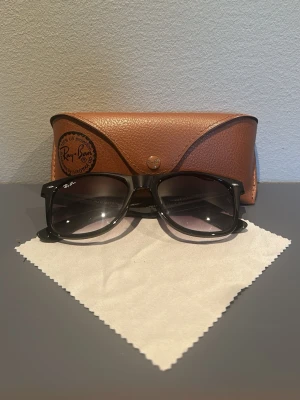 Svarta Ray-Ban Wayfarer solglasögon - Klassiska svarta Ray-Ban Wayfarer solglasögon med bred båge och mörka glas med blå/lila toning. Kommer med original brunt fodral och putsduk. Perfekta för dig som vill ha en tidlös och ikonisk look.