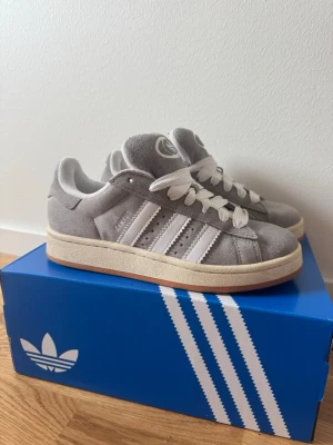 Adidas Campus 00 - Nästan oanvända, säljes eftersom jag har fått större fötter och inte längre kan ha storlek 38. Snygga grå Adidas Campus 00 sneakers. Skorna har breda vita skosnören och Adidas-logga på plösen. Perfekta för en clean och stilren look.