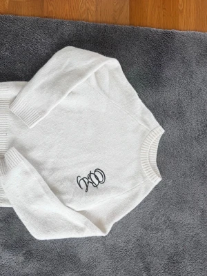 Vit stickad tröja med broderad text - Säljer en vit stickad tröja med lång ärm och ribbade muddar. Tröjan har en broderad svart textdetalj på framsidan och rund halsringning. Perfekt för dig som gillar enkel men snygg stil. Lite oversized fit så passar nån storlek större.