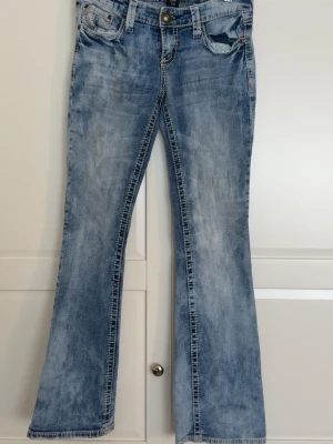 Ljusblå bootcut jeans med broderi - Säljer ett par ljusblå bootcut jeans som är låga i midjan med slitningar och snygga broderade detaljer på bakfickorna. Jeansen har klassisk femficksmodell, knappgylf och är tillverkade i denim. Perfekta för dig som gillar en avslappnad men trendig stil.