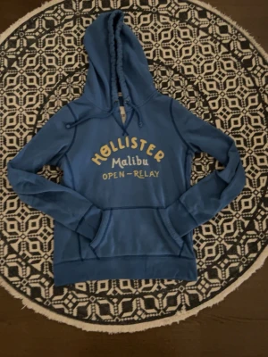 Blå hoodie från Hollister Malibu - Blå hoodie från Hollister med broderad gul och vit text 'Hollister Malibu Open-Relay' på bröstet. Klassisk känguruficka, dragsko i huvan och mjukt material. Perfekt för en avslappnad och cool stil.