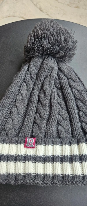 Grå kabelstickad mössa med tofs - Säljer en grå kabelstickad mössa med stor tofs på toppen och ribbad kant i vitt och grått. Mössan har en liten patch med bokstaven R framtill. Perfekt för kalla dagar och riktigt snygg till vinterlooken.