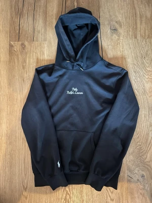 Svart hoodie från Polo Ralph Lauren - Svart hoodie med broderad Polo Ralph Lauren-logga på bröstet och liten vit polospelare på ärmen. Klassisk känguruficka framtill, justerbar huva och ribbade muddar. Perfekt för en avslappnad och stilren look.