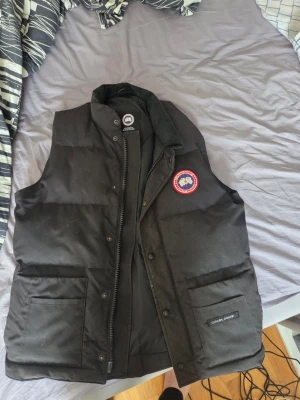 Svart dunväst från Canada Goose - Säljer en svart dunväst från Canada Goose med klassisk logga på bröstet och hög krage. Västen har två stora fickor framtill och stängs med både dragkedja och tryckknappar. Perfekt för lager-på-lager och riktigt varm under kalla dagar.