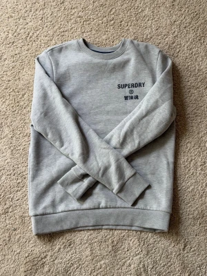 Grå sweatshirt från Superdry - Snygg ljusgrå sweatshirt från Superdry med rund halsringning och svart logga på bröstet. Tröjan har ribbade muddar vid ärmslut och nederkant. Perfekt för en avslappnad och stilren look.