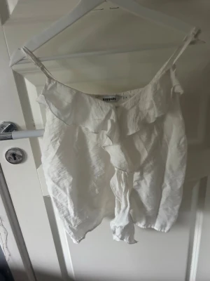 Vit linnetopp med volang från Kappahl - Söt vit linnetopp från Kappahl med tunna axelband och volangdetalj framtill. Toppen har knyt i nederkant och är perfekt för varma dagar. Lätt och luftig känsla som passar till jeans eller kjol.