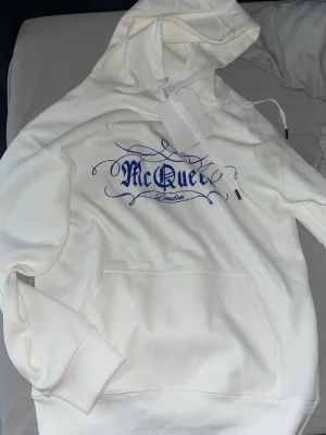 Vit hoodie från Alexander McQueen - Säljer en vit hoodie från Alexander McQueen med blått tryck på bröstet där det står 'McQueen London' i snygg stil. Hoodien har huva med dragsko och en stor magficka. Perfekt för dig som gillar streetwear och vill sticka ut lite extra.
