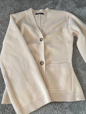 Beige kofta från Gina Tricot - Stilren beige kofta från Gina Tricot med två knappar framtill och en praktisk ficka på ena sidan. Koftan har ribbade muddar vid ärmslut och nederkant samt en djup V-ringning. Perfekt att slänga över en topp för en enkel och snygg look.