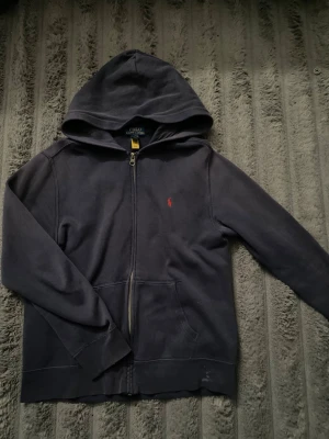 Mörkblå hoodie med dragkedja Polo - Mörkblå hoodie från Polo Ralph Lauren med dragkedja framtill och klassisk röd logga broderad på bröstet. Tröjan har huva, fickor på sidorna och ribbade muddar. Perfekt för en avslappnad och stilren look.