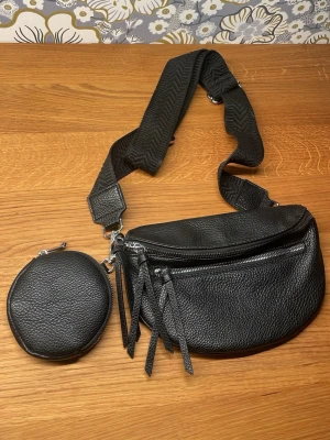 Svart crossbody handväska med myntficka - Snygg svart handväska i crossbody-modell med justerbar bred axelrem och matchande rund myntficka. Väskan är gjord i strukturerat konstläder med silverfärgade dragkedjor och detaljer. Perfekt för dig som vill ha både stil och funktion i ett.