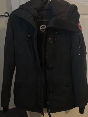 Svart dunjacka från Canada Goose - Säljer en svart dunjacka från Canada Goose med huva och dragkedja samt knappar framtill. Jackan har fickor med dragkedja och den klassiska röda logotypen på ärmen. Perfekt för kalla vinterdagar och riktigt varm.
