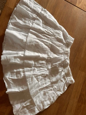 Vit volangkjol i bomull med inbyggda shorts - Söt vit kortkjol med volanger och resår i midjan. Kjolen är gjord i luftig bomull och har en avslappnad, flowy passform som passar perfekt till varma dagar. Enkel att matcha med olika toppar för en fräsch sommarlook. Med inbyggda shorts