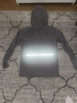 Grå vindjacka från Under Armour - Snygg grå vindjacka från Under Armour med huva och hel dragkedja framtill. Jackan har reflekterande detaljer och logga både fram och bak, samt två fickor med dragkedja. Perfekt för utomhusaktiviteter och träning.