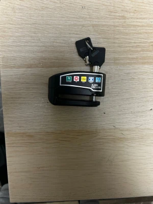 Svart bromsskivelås med larm och nycklar - Säljer ett svart bromsskivelås med inbyggt larm och två nycklar. Låset har tydliga symboler och texten 'ALARM DISC LOCK' på sidan. Perfekt för att säkra din moped eller motorcykel mot stöld.