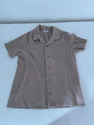 Beige kortärmad skjorta med knappar - Snygg beige skjorta i ribbad bomull med korta ärmar och klassisk krage. Skjortan har en bröstficka och stängs med knappar framtill. Perfekt för en avslappnad och stilren look.