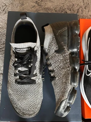 Nike Vapormax grå sneakers - Säljer ett par Nike Vapormax sneakers i grått med svart sula och svarta detaljer. Ovandelen är stickad i mesh för extra komfort och ventilation. Skorna har en tydlig logga på sidan och en unik, genomskinlig sula som ger en futuristisk look.