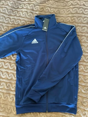 Blå träningsjacka från Adidas - Snygg blå träningsjacka från Adidas med vit logga på bröstet och vita detaljer längs axlarna. Jackan har hög krage och dragkedja framtill, perfekt för träning eller sportiga outfits.
