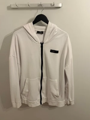 Vit hoodie med dragkedja från Plein Sport - Snygg vit hoodie från Plein Sport med svart dragkedja och två stora fickor framtill. Hoodien har en klassisk huva och en diskret logga på bröstet. Perfekt för en avslappnad och sportig stil.