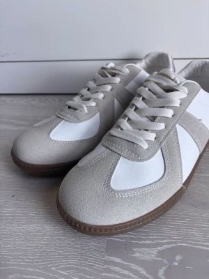 Vita o beiga MM Gats  - Snygga MM Gats i vitt och beige med ovandel i mocka och släta partier. Skorna har vita snören och en klassisk gummisula i brunt. Perfekta för dig som gillar stilrena och tidlösa sneakers med en modern touch.