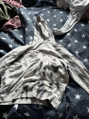 Grå tie-dye hoodie med huva - Säljer en grå och svart tie-dye hoodie med huva. Hoodien har en avslappnad passform och ribbade muddar vid ärmslut och nederkant. Perfekt för dig som gillar streetwear och vill ha en unik look.