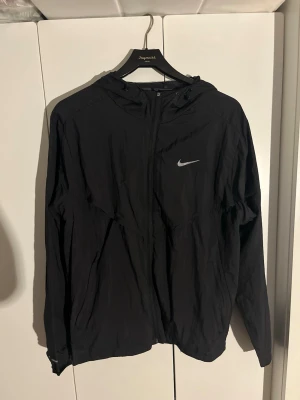 Nike Miller svart - Svart vindjacka från Nike med dragkedja och huva. Jackan har en diskret Nike-logga på bröstet och är tillverkad i ett lätt, vindtätt material. Perfekt för blåsiga dagar och sportiga outfits.
