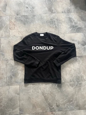 Svart sweatshirt från Dondup - Säljer en svart sweatshirt från Dondup med vit logga tryckt över bröstet. Tröjan har rund halsringning, långa ärmar och ribbade muddar vid ärmslut och nederkant. Perfekt för en avslappnad och stilren look.