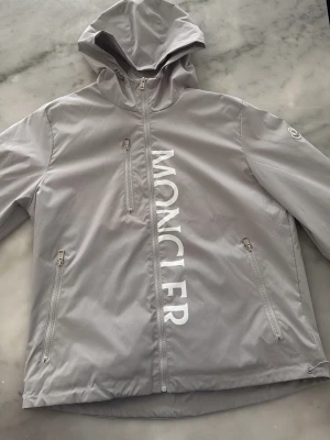 Grå vindjacka från Moncler - Snygg grå vindjacka från Moncler med huva och dragkedja framtill. Jackan har stora vita Moncler-bokstäver längs dragkedjan, tre fickor med dragkedja och Moncler-logga på ärmen. Tillverkad i ett lätt, glansigt material som står emot vind.