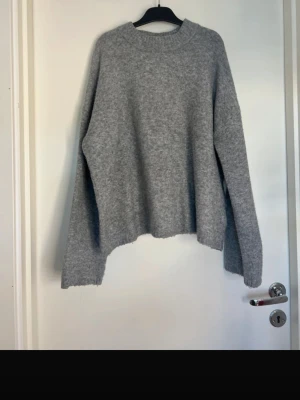 Grå stickad tröja från H&M - Mysig grå stickad tröja från H&M med rund halsringning och avslappnad passform. Tröjan har långa ärmar och ribbade muddar vid halsen. Perfekt för kyliga dagar och enkel att matcha med olika outfits.