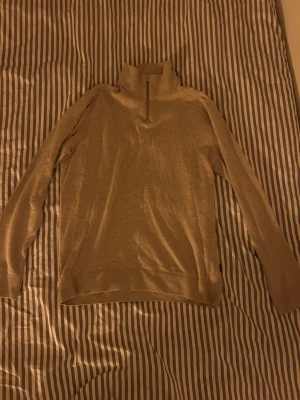 Beige stickad tröja med halv dragkedja - Beige stickad tröja med hög krage och halv dragkedja framtill. Tröjan har långa ärmar och ribbstickade muddar vid ärmslut och nederkant. Perfekt för lager-på-lager och en stilren look.