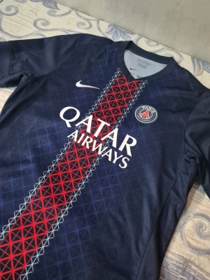 PSG matchtröja - Snygg PSG matchtröja från Nike i mörkblått med röd och vit grafisk rand mitt på bröstet. Tröjan har klubbmärke, Qatar Airways-tryck och Dri-FIT-material som andas. Perfekt för fotboll eller streetwear.
