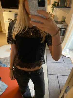 Svart croppad spetsblus med leopardmönster - Säljer en svart, croppad blus i genomskinlig spets med leopardmönster. Toppen har korta ärmar och vågig kant nertill för en extra snygg detalj. Har ett linne under på de två första bilderna
