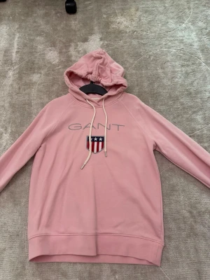 Rosa hoodie från GANT med logga - Säljer en ljusrosa hoodie från GANT med broderad logga och sköld på bröstet. Hoodien har dragsko i huvan och långa ärmar. Tillverkad i mjukt material som känns skönt mot huden. Perfekt för en avslappnad och trendig look.