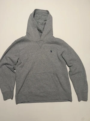 Grå hoodie från Polo Ralph Lauren - Klassisk grå hoodie från Polo Ralph Lauren med en svart broderad logga på bröstet. Hoodien har känguruficka framtill, och mjukt bomull på insidan. 