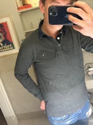 Mörkgrå långärmad från gant  - Snygg mörkgrå långärmad gant med mörkblå krage och knappar framtill. Skjortan har en liten broderad detalj på bröstet och är tillverkad i mjukt bomullsmaterial. Perfekt för en avslappnad men stilren look.