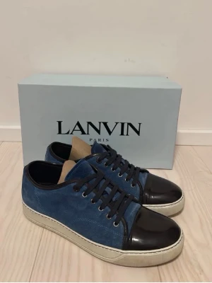 Blå Lanvin - Snygga blå Lanvin med svart lackad tå och matchande svarta skosnören. Ovandelen är i blå mocka och sulan är vit med en klassisk siluett. Perfekta för dig som vill ha en stilren och exklusiv look.