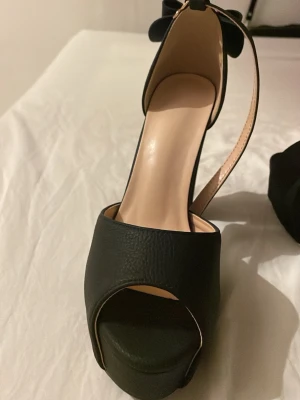 Svarta pumps med rosett och platå - Snygga svarta pumps med hög klack och platåsula. Skorna har en öppen tå och en elegant rosett baktill vid hälen. Tillverkade i ett material som liknar läder och med en smal ankelrem för extra stöd.