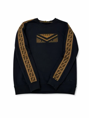Mörkblå Fendi sweatshirt med logoband - Snygg mörkblå sweatshirt från Fendi med bruna logoband längs ärmarna och ett stort Fendi-emblem på bröstet. Tröjan har rund halsringning, ribbade muddar och är tillverkad i mjukt material. Perfekt för dig som gillar streetwear och exklusiva detaljer.