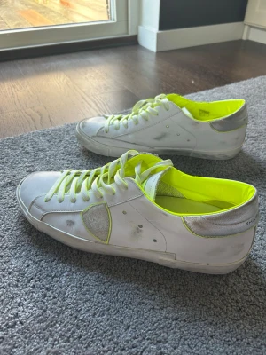 Vita sneakers Philippe Model  - Säljer ett par vita Philippe Model sneakers med neon-gula detaljer och innersula. Skorna har silverfärgade partier vid hälen och klassisk låg siluett. Tillverkade i läder med snygga kontrastfärger och ikonisk sköld på sidan.