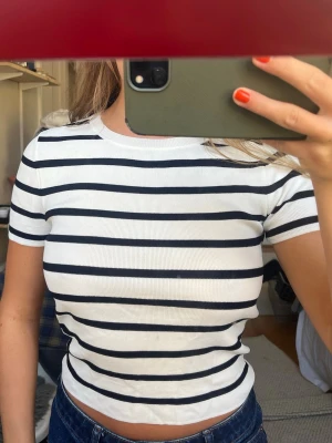Randig vit och svart croppad topp - Säljer en vit croppad topp med svarta horisontella ränder. T-shirten har rund halsringning och korta ärmar. Materialet är ribbat och stretchigt, vilket ger en bekväm passform. Perfekt till jeans eller kjol för en enkel och snygg look.