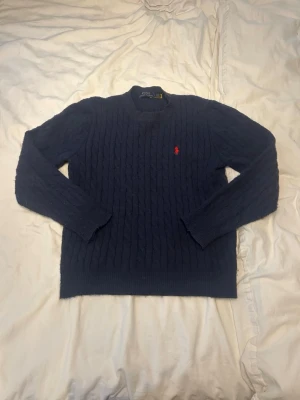 Mörkblå kabelstickad tröja Polo Ralph Lauren - Mörkblå kabelstickad tröja från Polo Ralph Lauren med rund halsringning och den klassiska röda logotypen broderad på bröstet. Tröjan har långa ärmar och ribbade muddar vid ärmslut och nederkant. Perfekt för lager på lager och stilren look.Den är väl använd med en del små noppor.Jag skulle att den är för personer som är runt 160