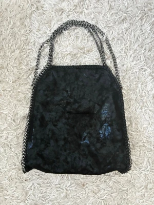 Svart axelväska med kedjedetaljer - Snygg svart axelväska i mjukt, glansigt material med silverfärgade kedjor som handtag och längs kanterna. Väskan har en rymlig form och passar perfekt för dig som vill ha plats för allt du behöver. Kedjorna ger en cool och trendig look.