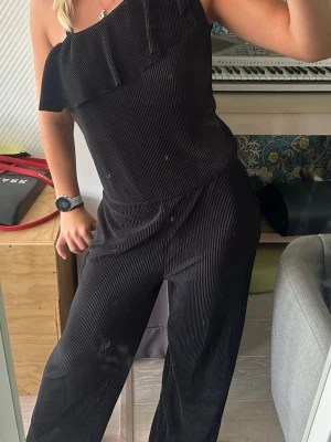 Svart plisserad byxdress med volang - Säljer en svart plisserad jumpsuit med en axelband och volangdetalj över bröstet. Vida ben och mjukt flowy tyg, så himla skön!!! Storlek 158/164 men har funkat super på mig som är 167 och en vanlig S för den är väldigt stretchig 🥰