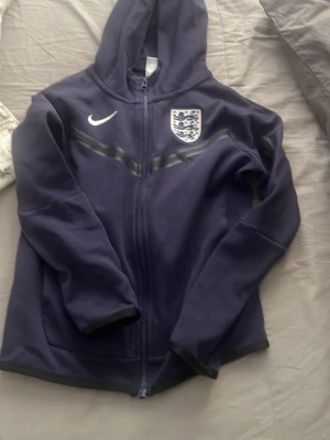 Mörkblå England Nike tech  - Snygg mörkblå hoodie med dragkedja från Nike, prydd med Englands landslagsmärke på bröstet. Tröjan har huva, två fickor och en diskret grå detalj över bröstet. Perfekt för fotbollsfans och sportiga dagar.