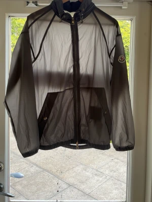 Grå vindjacka från Moncler - Snygg grå vindjacka från Moncler med dragkedja framtill, två snedställda fickor med knapp och elastiska muddar. Jackan har en lätt och tunn design, perfekt för blåsiga dagar. Moncler-logga på vänster ärm ger en exklusiv touch.