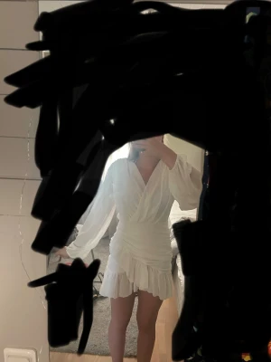 Vit omlottklänning med volang - Kort vit klänning med omlott-design och v-ringning. Klänningen har långa ärmar och volangdetaljer vid nederkanten. Perfekt för dig som gillar en feminin och trendig stil.