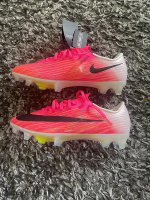 Nike rosa Mercurial fotbollsskor - Säljer ett par Nike Mercurial fotbollsskor i en grymt snygg rosa färg med svart swoosh. Skorna har en lätt ovandel i syntetmaterial och genomskinlig sula med Air Zoom-dämpning och inslag av gult och blått. Perfekta för dig som vill ha max speed på planen.