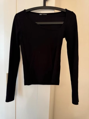 Svart långärmad topp från Zara - En stilren svart långärmad topp från Zara med en bred, något asymmetrisk ringning. Toppen har en slim passform och är tillverkad i ett mjukt material som sitter snyggt på kroppen. Perfekt att matcha med jeans eller kjol.